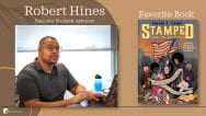Portrait of Robert Hines, Student Success Advisor, at their desk. Their favorite book, Stamped from the Beginning: A Graphic History of Racist Ideas in America, is pictured next to their portrait.